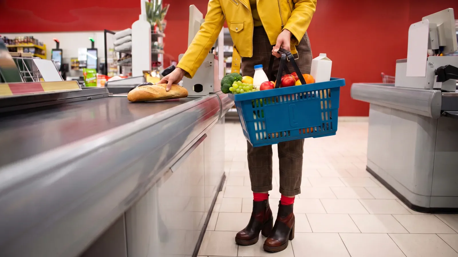 Mulher de jaqueta amarela diante do caixa de um supermercado, com a cesta azul esperto